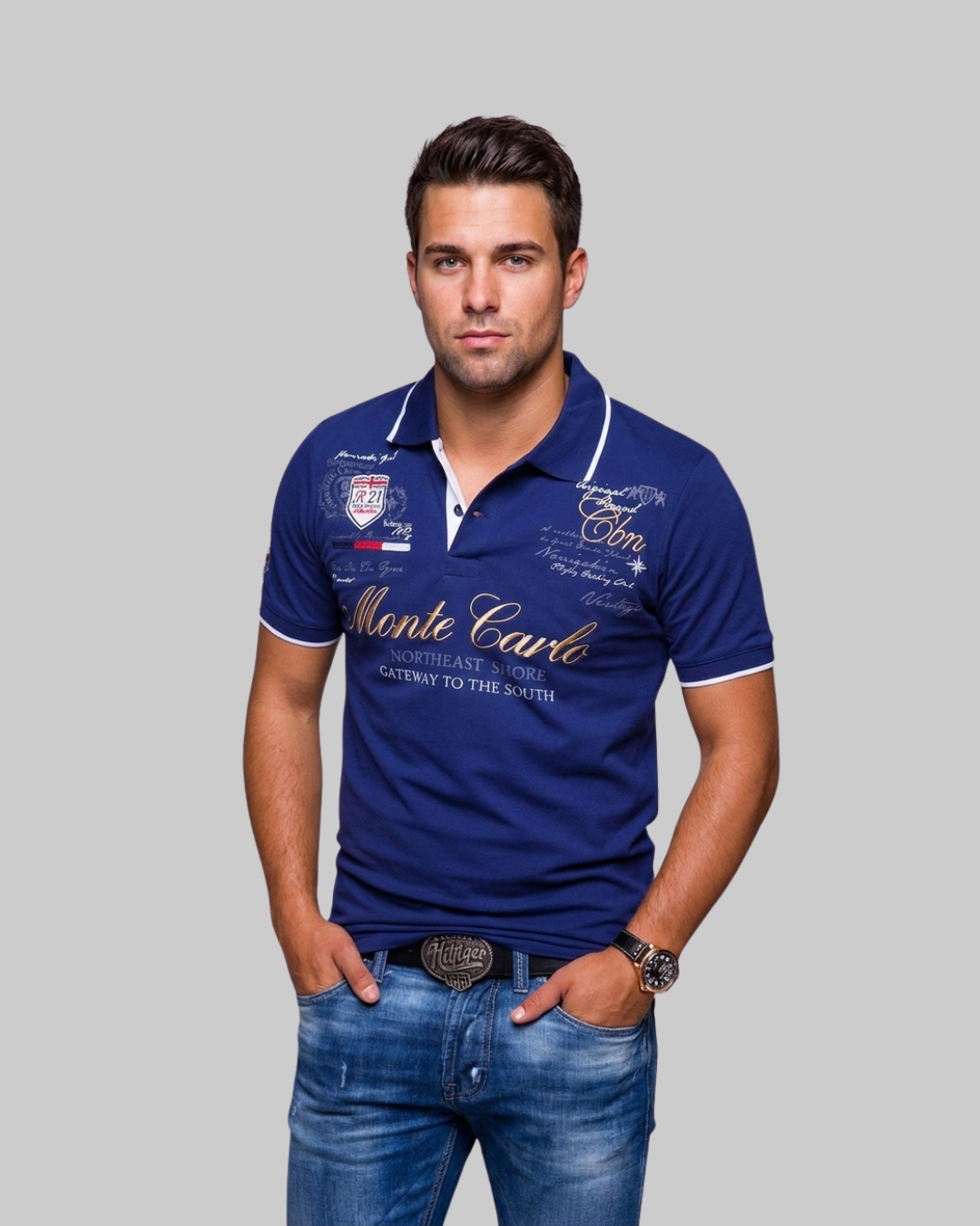 A man wearing a navy blue polo shirt with white and gold graphic details, paired with blue jeans, standing against a solid gray background.