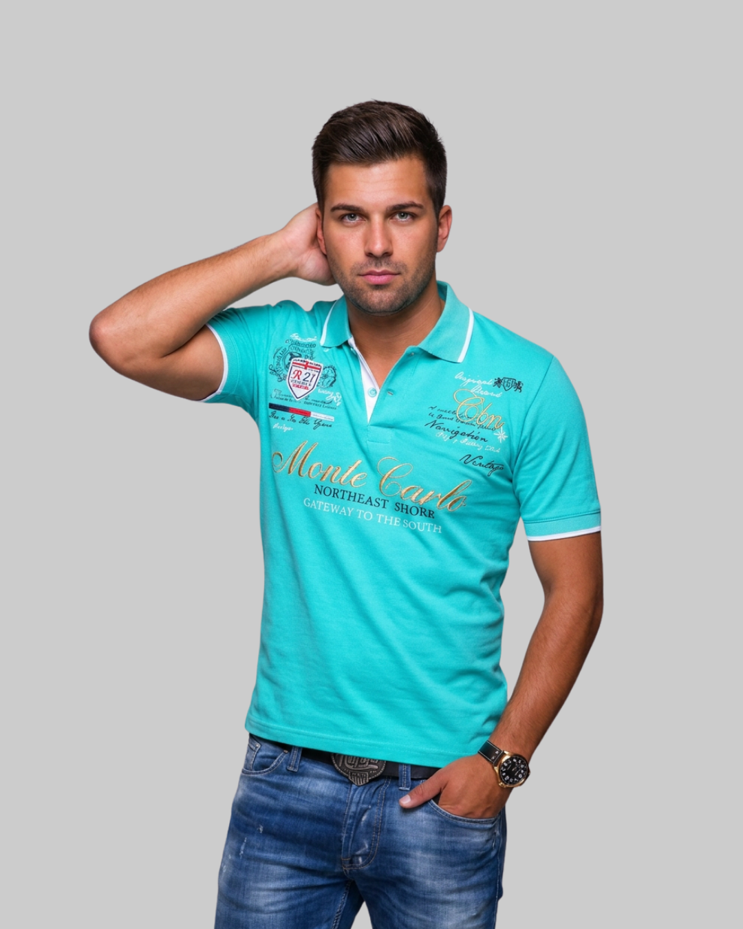 A man modeling a turquoise blue polo shirt with white trim and gold script lettering, paired with blue jeans, against a solid gray background.
