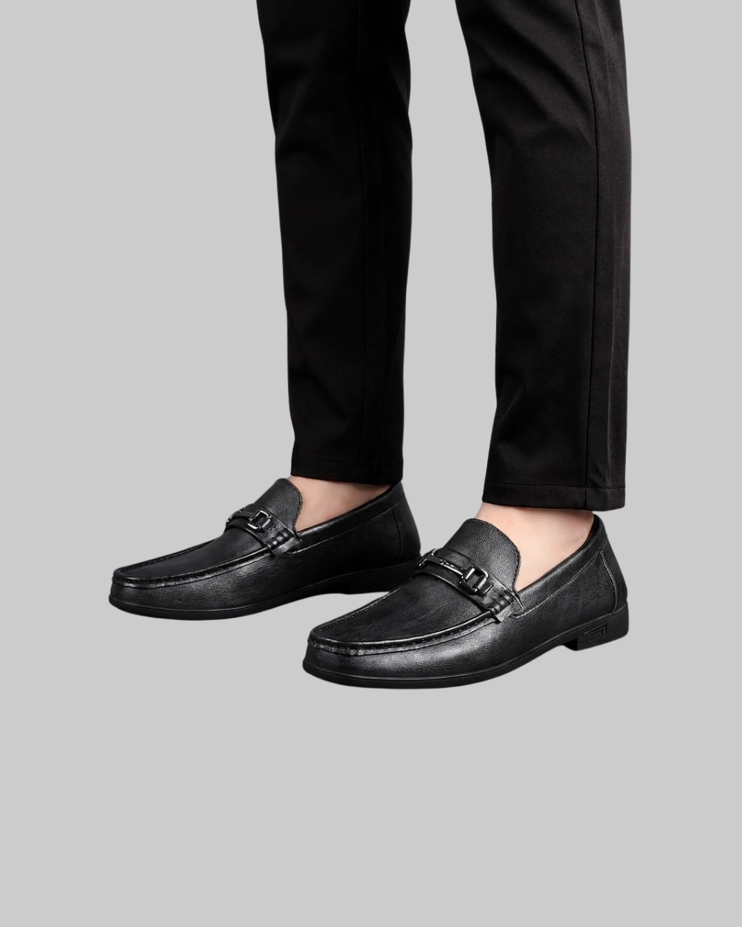 Black leather loafers with a small buckle detail on the vamp, shown on a model wearing black trousers against a solid gray background.