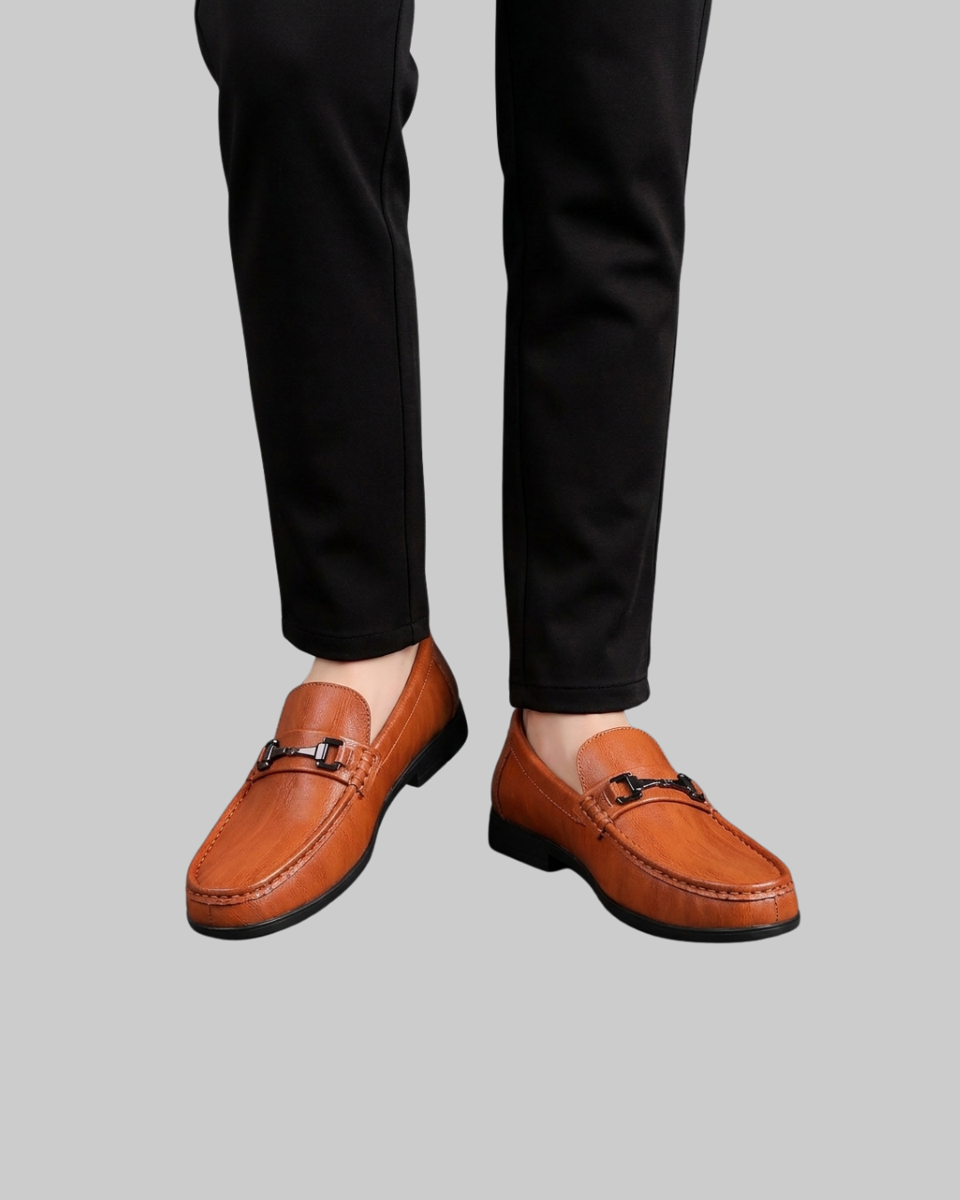 A person wearing slim black trousers and tan leather penny loafers with black hardware, shown from the calves down against a plain light gray background.