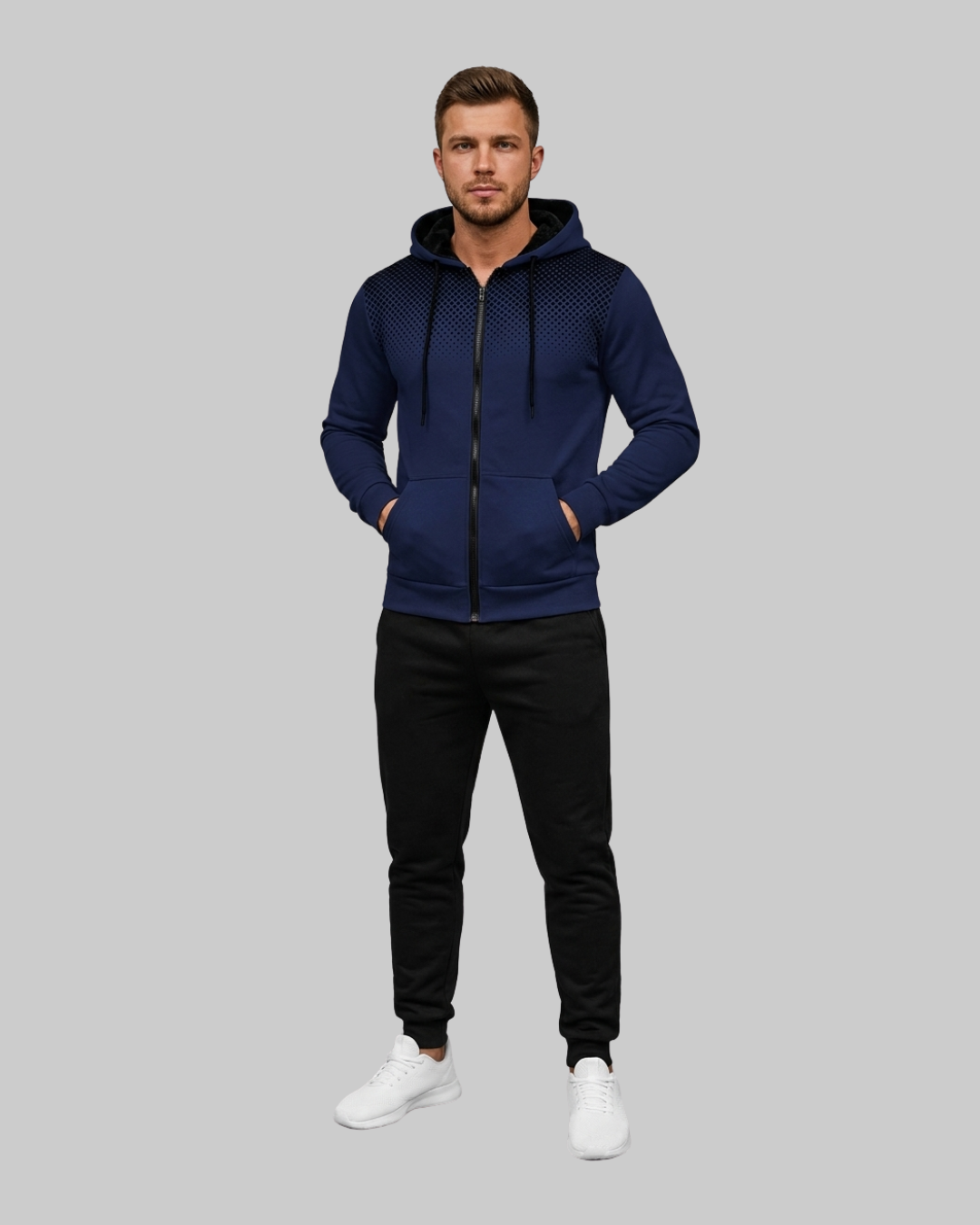 A man modeling a navy blue zip-up hoodie with a textured pattern on the chest, black pants, and white sneakers against a solid gray background.