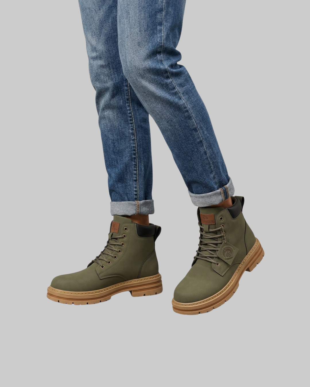 A pair of olive green lace-up boots with tan soles, shown on a model wearing blue jeans against a solid gray background.