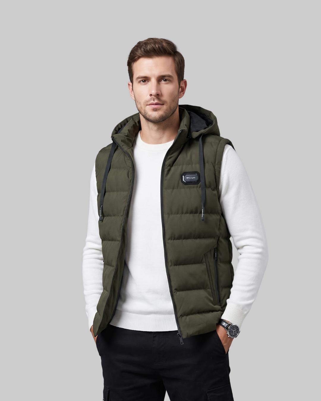 A man wearing an olive green hooded puffer vest over a white long-sleeve shirt and dark pants, standing against a solid gray studio background.