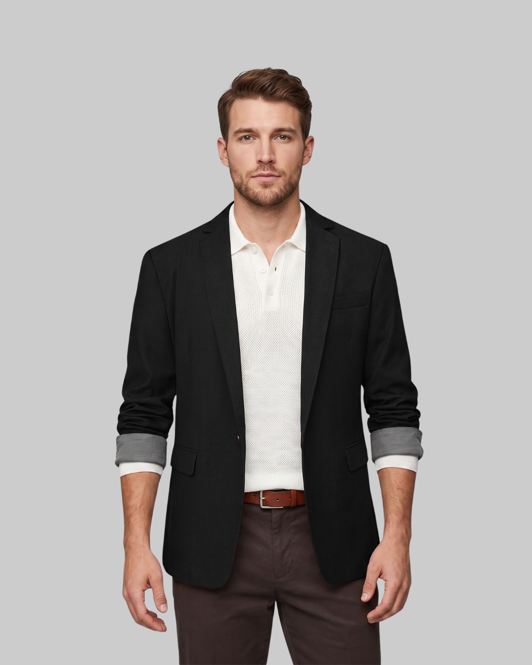 A man wearing a black blazer over a white polo shirt, brown trousers, and a brown belt, standing against a solid gray background.