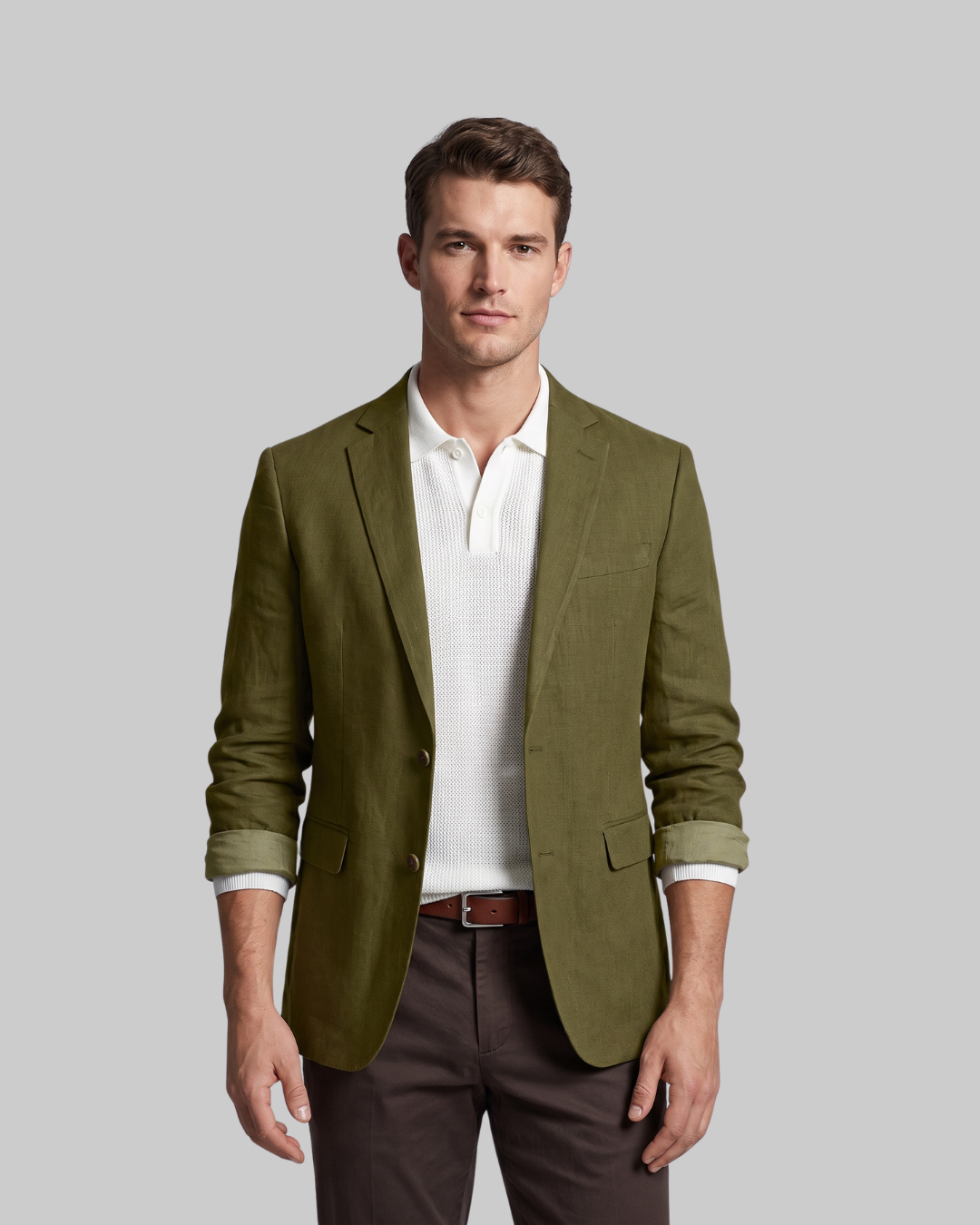 A man wearing an olive green blazer over a white polo shirt and dark brown trousers, standing against a solid gray background.