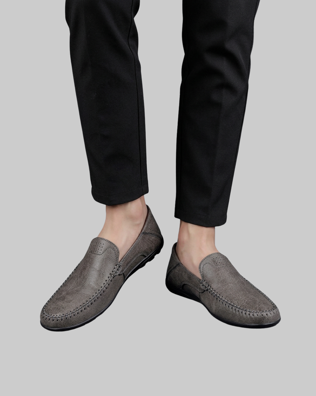 A person wearing black trousers and gray slip-on loafers with decorative stitching, shown from the calves down against a plain light gray background.