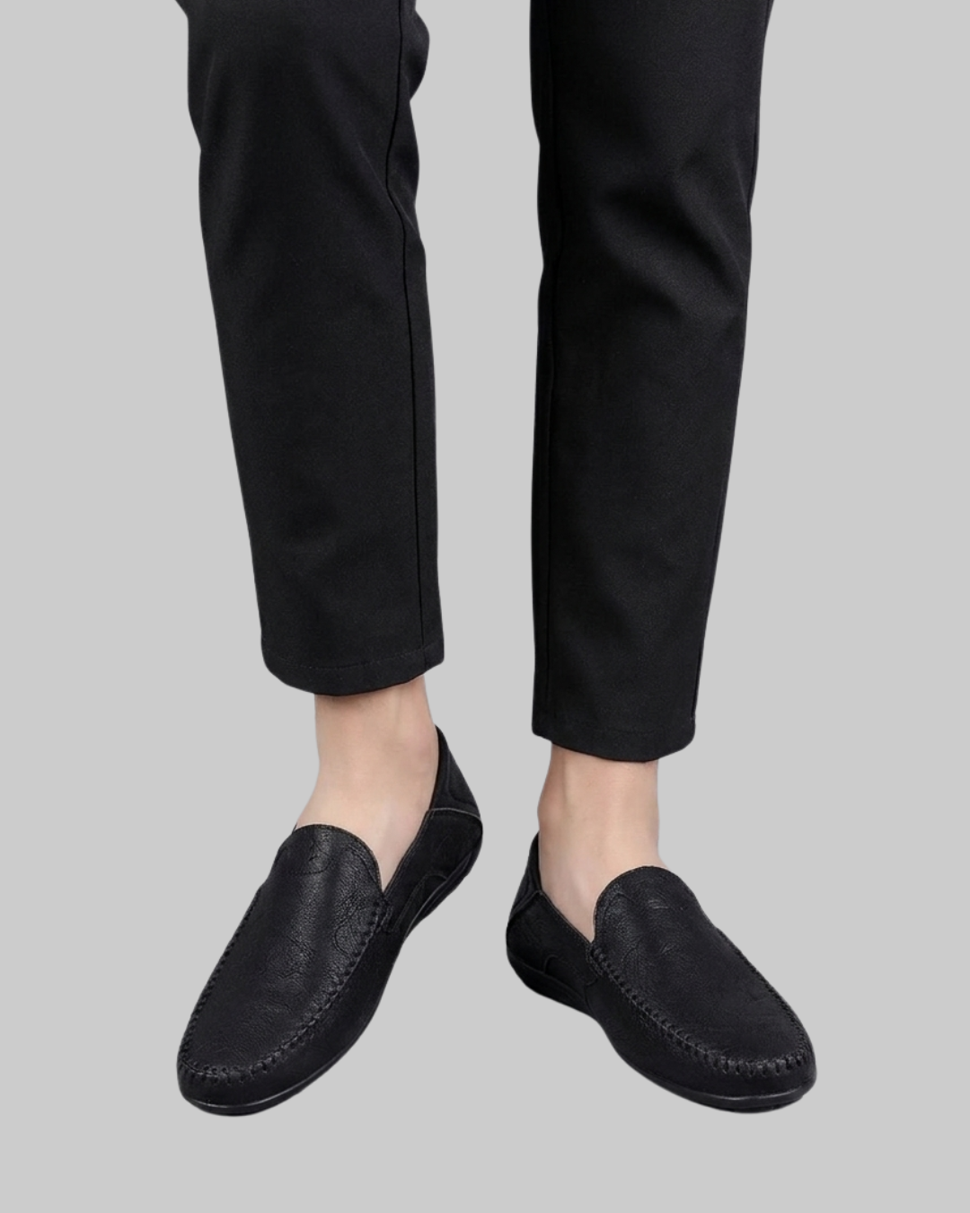 Black leather slip-on loafers worn with black trousers, shown from the feet up against a solid gray background.