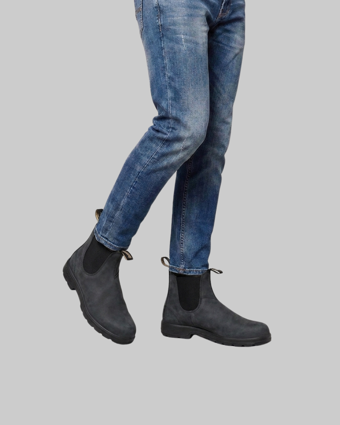 A person wearing blue jeans and dark gray Chelsea-style ankle boots, shown from the knees down against a plain light gray background.