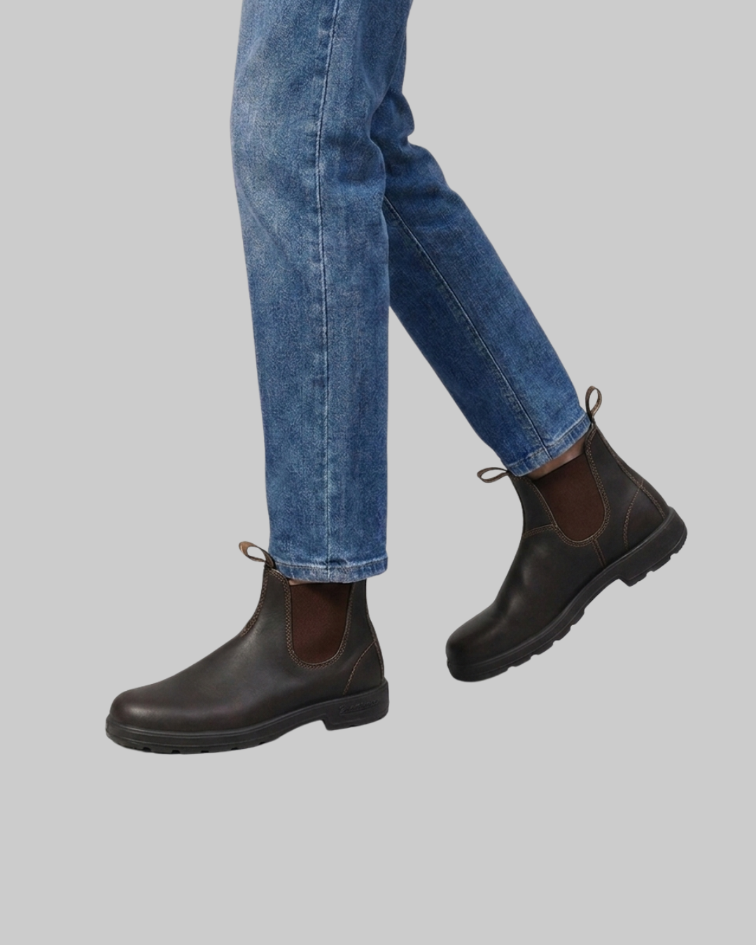 Dark brown Chelsea-style ankle boots worn with blue jeans against a solid gray background.
