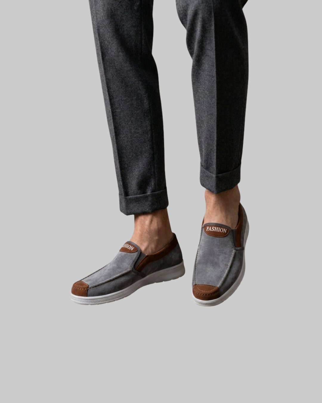 A person wearing gray slip-on loafers with brown accents and dark charcoal pants, shown from the calves down against a plain light gray background.