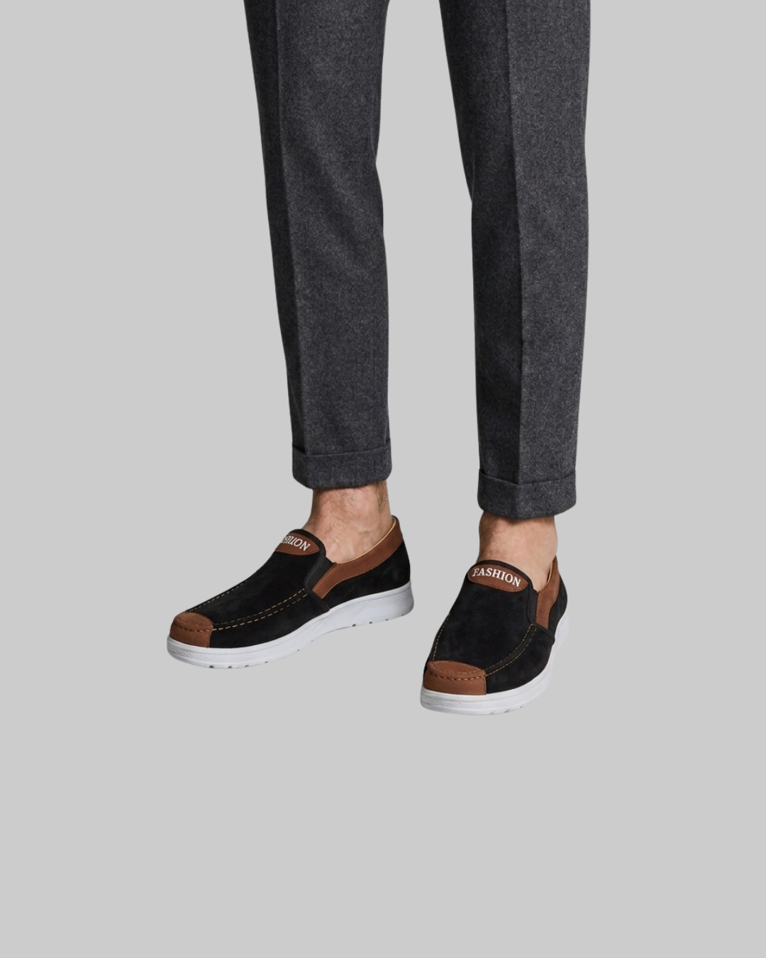 A person wearing dark charcoal gray trousers and black slip-on shoes with brown accents and white soles, shown from the calves down against a plain light gray background.