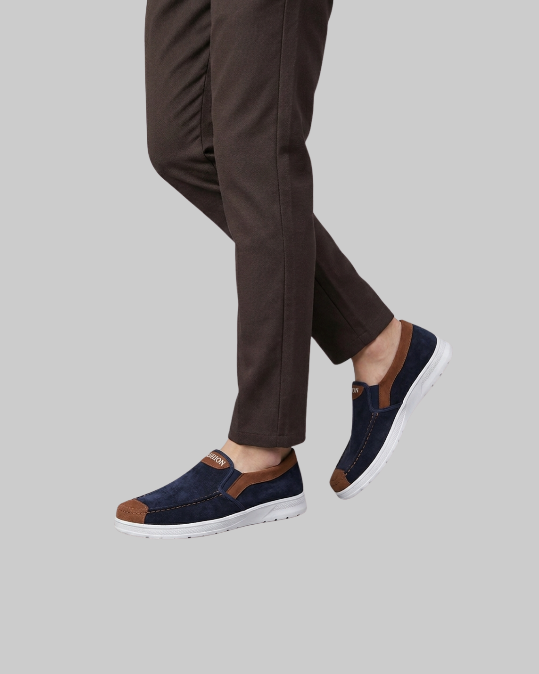 A person wearing dark brown trousers and navy slip-on sneakers with brown accents and white soles, shown from the calves down against a plain light gray background.