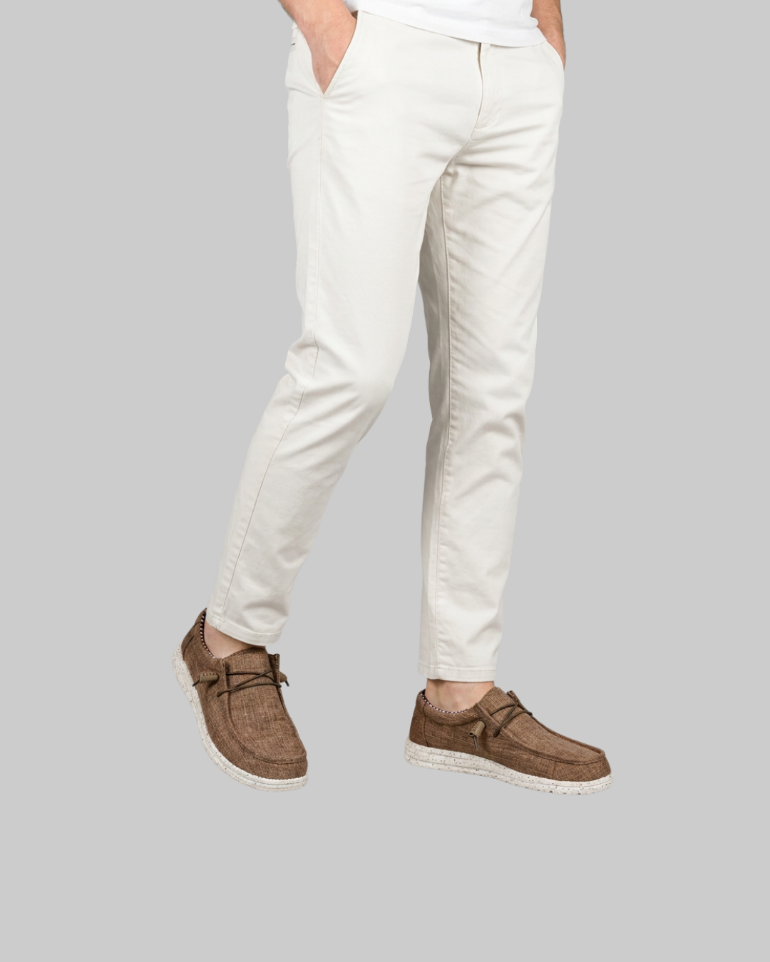 A person wearing slim-fit white chino pants and brown suede boat shoes, shown from the waist down against a solid gray background.