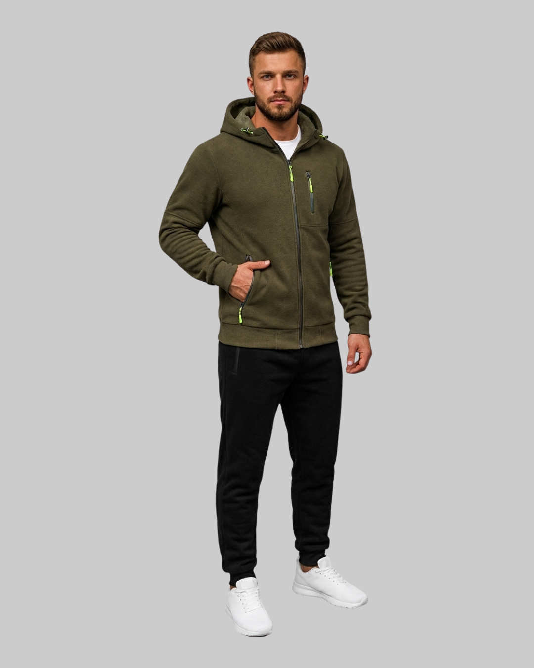 A man modeling an olive green zip-up hoodie with neon green accents, paired with black sweatpants and white sneakers, against a solid gray background.