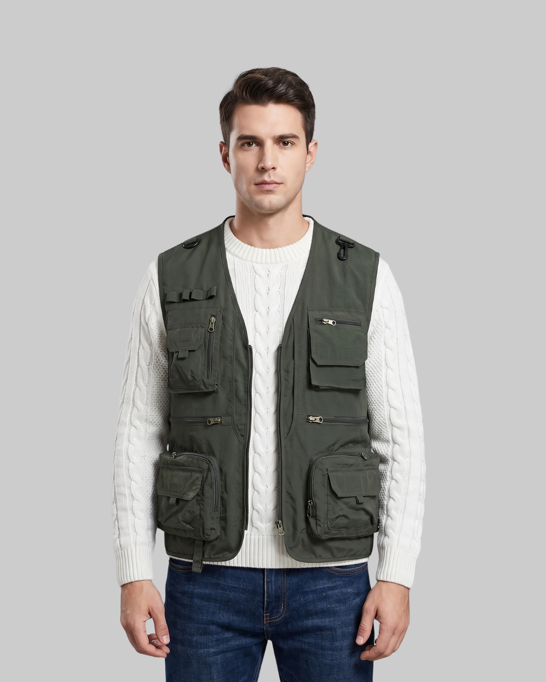 A man wearing an olive green utility vest over a white cable-knit sweater and blue jeans, standing against a solid gray background.