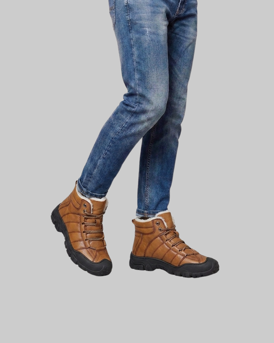 A person wearing brown leather lace-up ankle boots with black soles and blue jeans, shown from the knees down against a solid gray background.