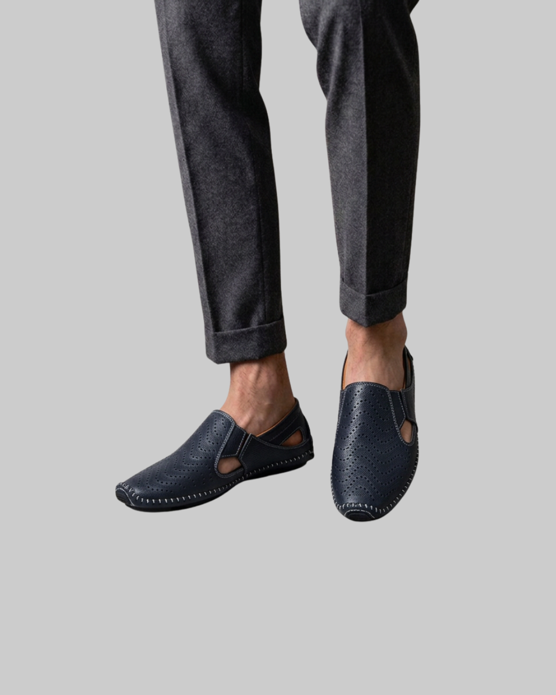 A person wearing dark navy slip-on leather shoes with cutout details and contrast stitching, paired with dark gray trousers, shown against a solid light gray background.