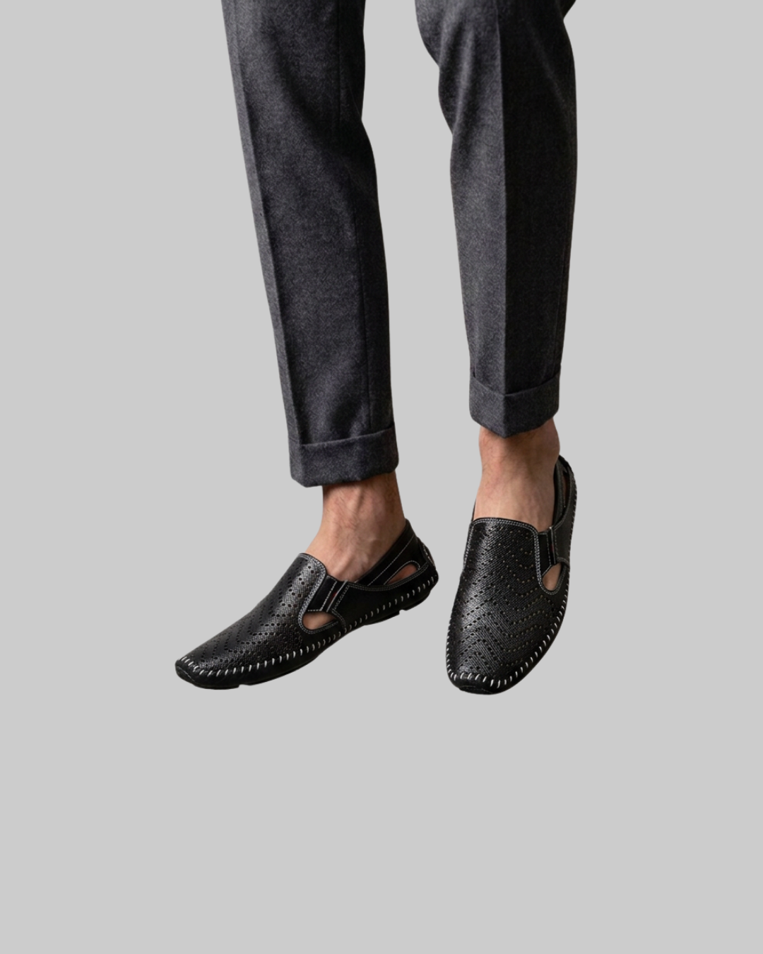 Black leather slip-on loafers with textured grain and decorative stitching, shown on a model wearing dark gray trousers against a solid gray background.