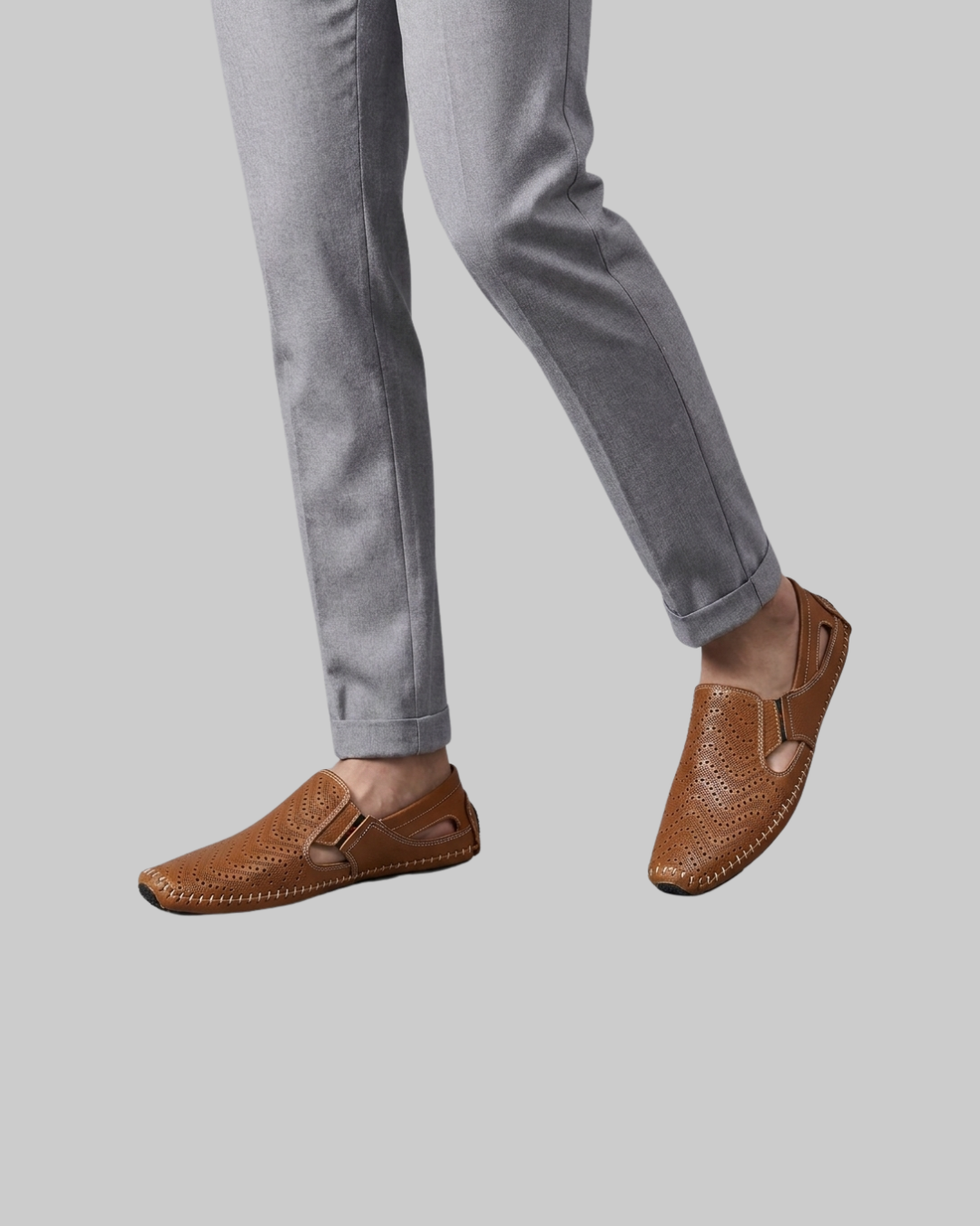 A person wearing light gray pants and brown leather slip-on loafers with perforated detailing, shown against a plain light gray background.