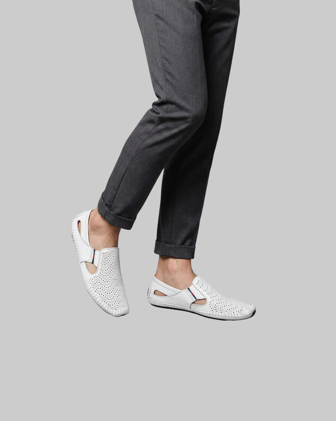 White perforated slip-on sandals worn with dark gray trousers, shown from the side against a solid gray background.
