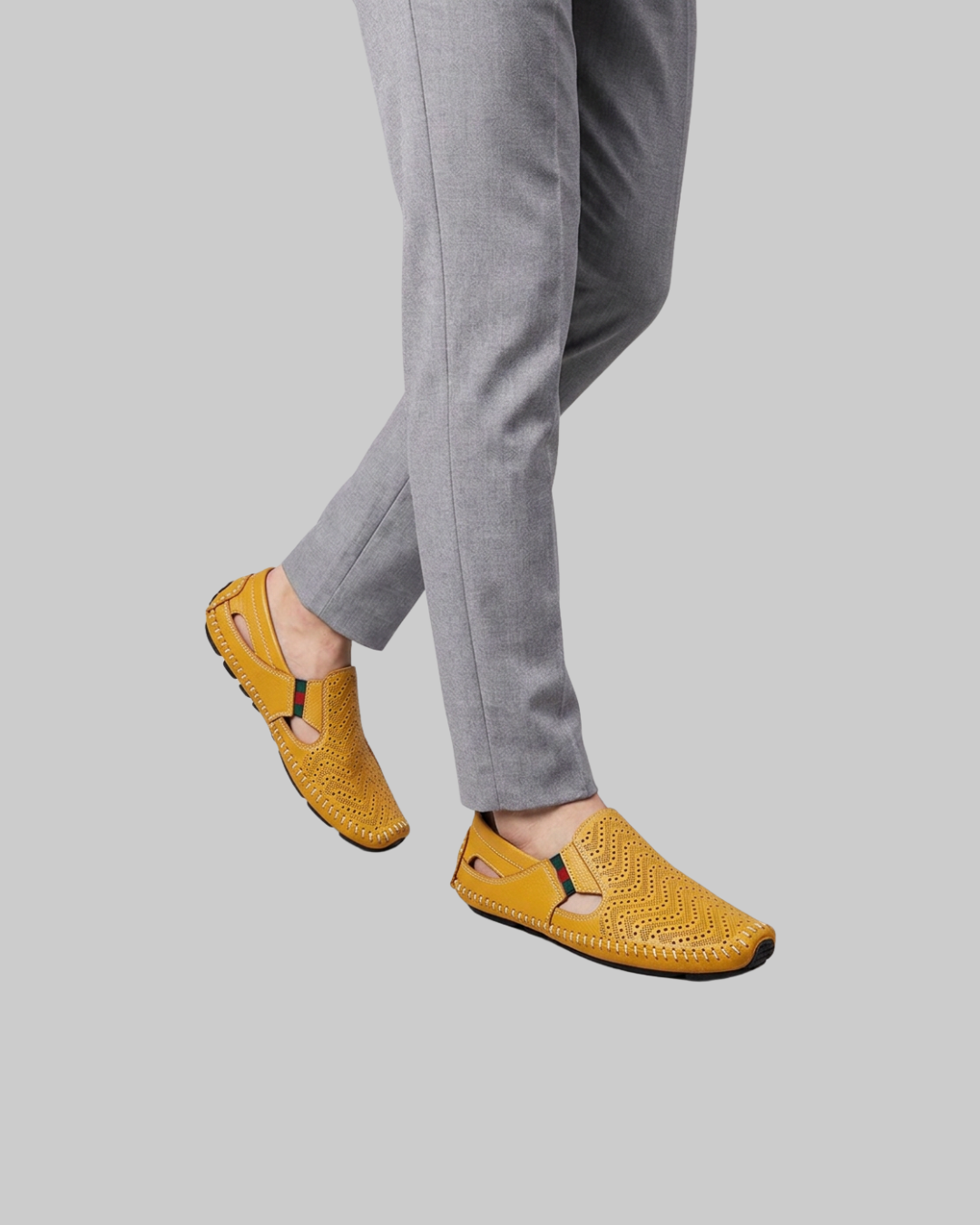 A person wearing mustard yellow perforated slip-on loafers with gray trousers, shown from the knees down against a solid gray background.
