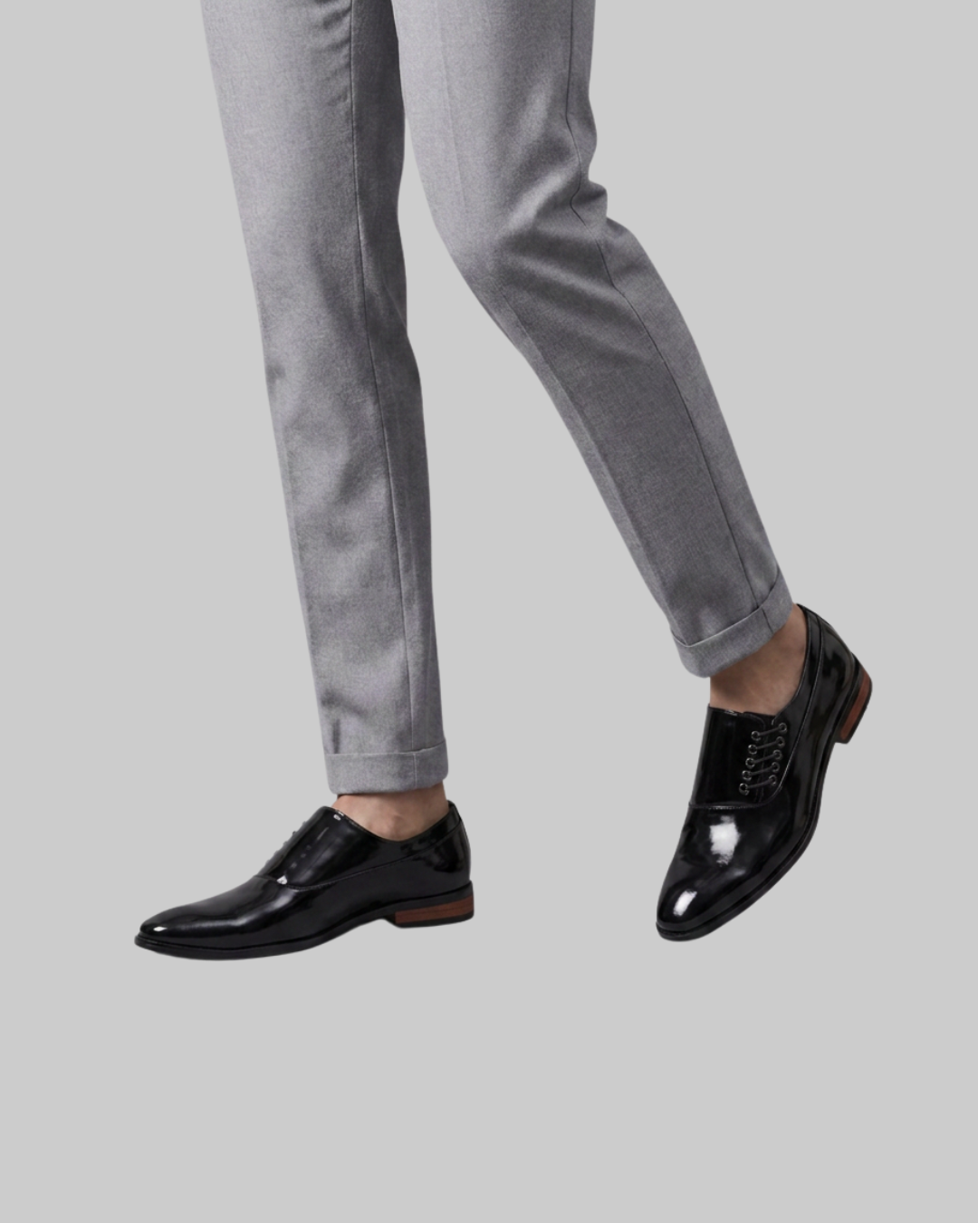 A person wearing slim-fit light gray trousers and glossy black lace-up dress shoes, shown from the calves down against a plain light gray background.