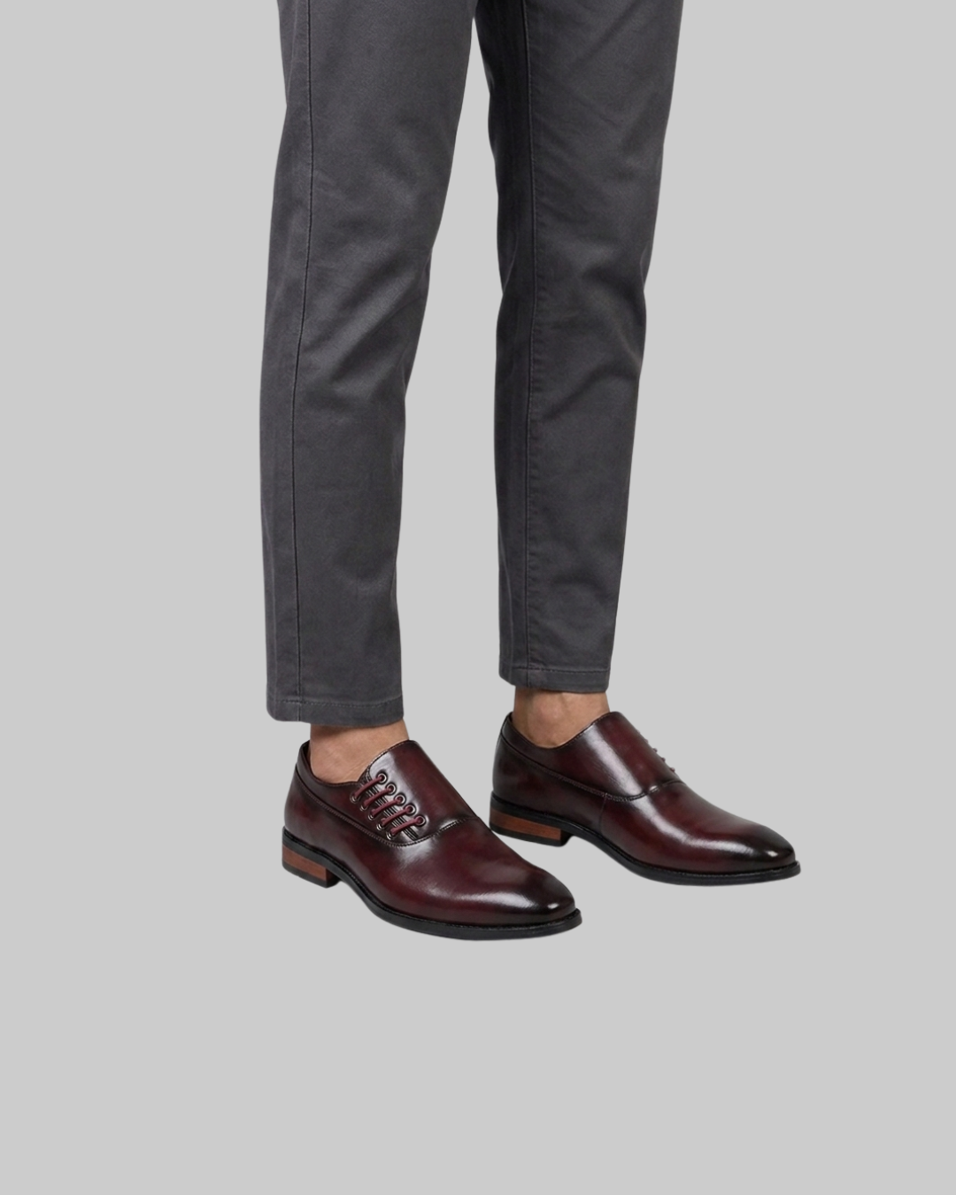 A pair of men's dark burgundy oxford shoes with a glossy finish, worn with dark gray trousers against a solid gray background.