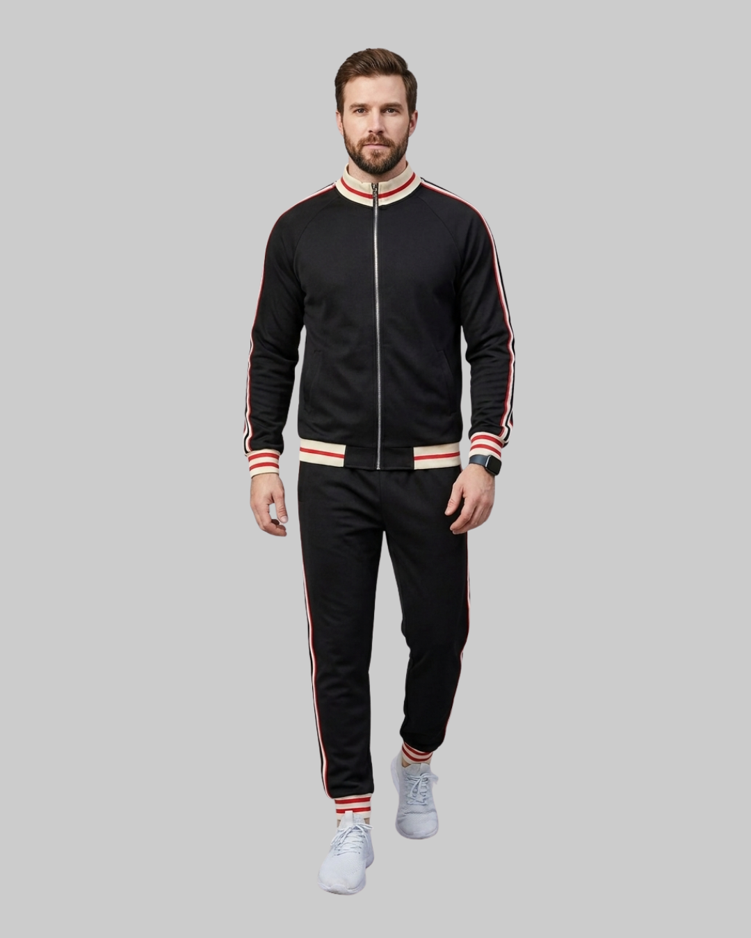 A man modeling a black zip-up track jacket and matching black track pants with red and white striped ribbed trim, paired with white sneakers, against a solid gray background.