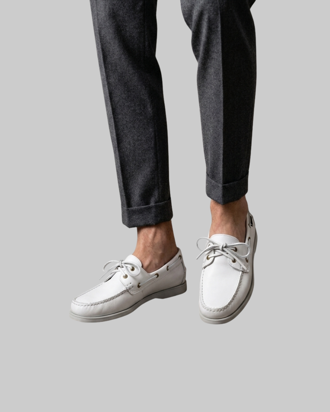 White leather boat shoes with white laces, shown on a model wearing dark gray trousers against a solid gray background.