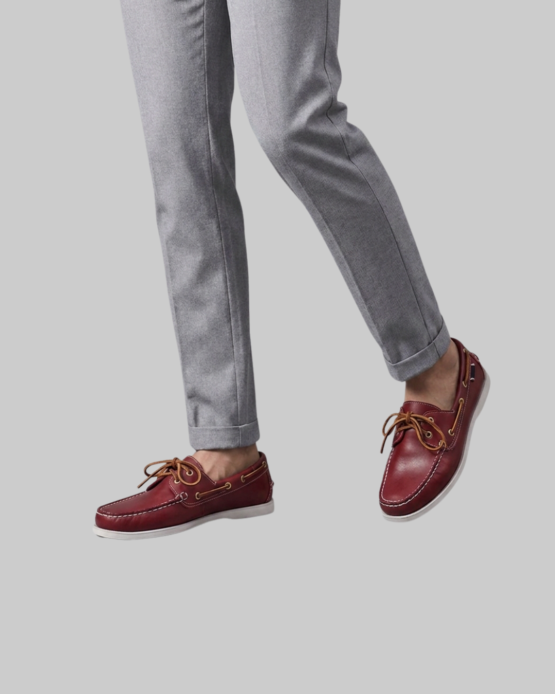 A person wearing gray pants and burgundy boat shoes with tan laces, shown from the knees down against a light gray background.