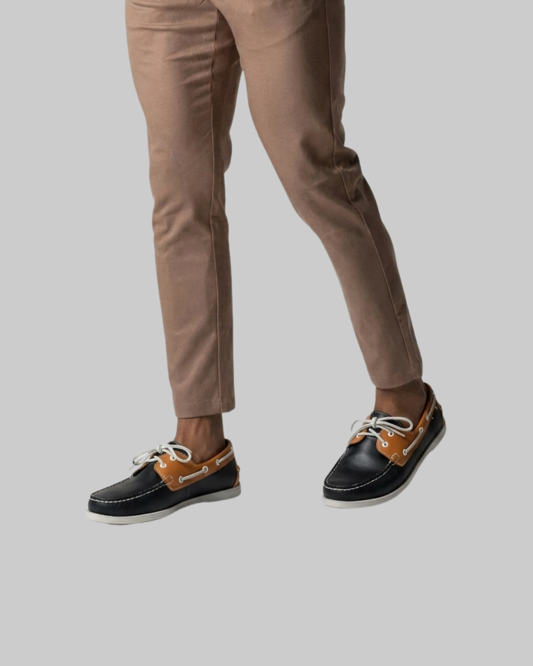 A person wearing slim tan pants and black boat shoes with tan side panels and white laces, shown from the waist down against a light gray studio background.