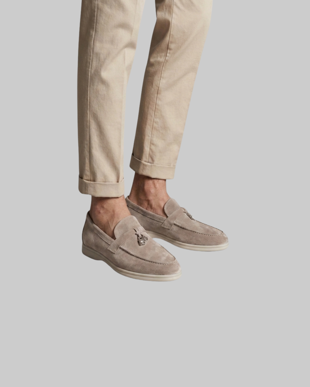 A person wearing beige trousers and taupe suede moccasin loafers with small metallic decorative details on the vamp, shown against a solid light gray background.
