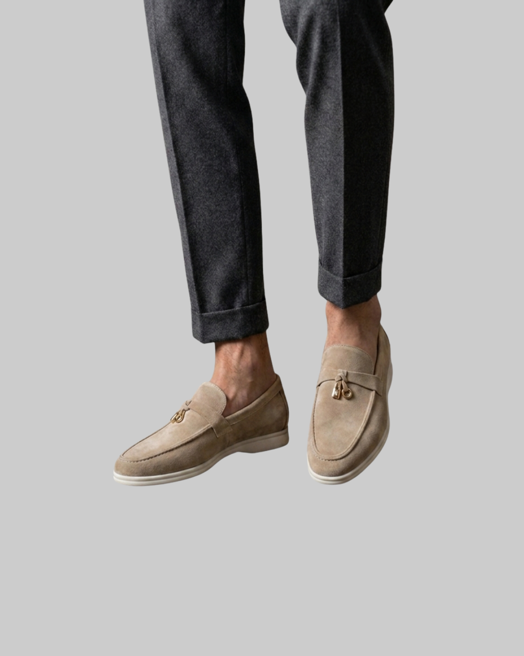 A person wearing dark charcoal gray trousers and light beige suede loafers with gold-tone logo hardware, shown from the calves down against a plain light gray background.