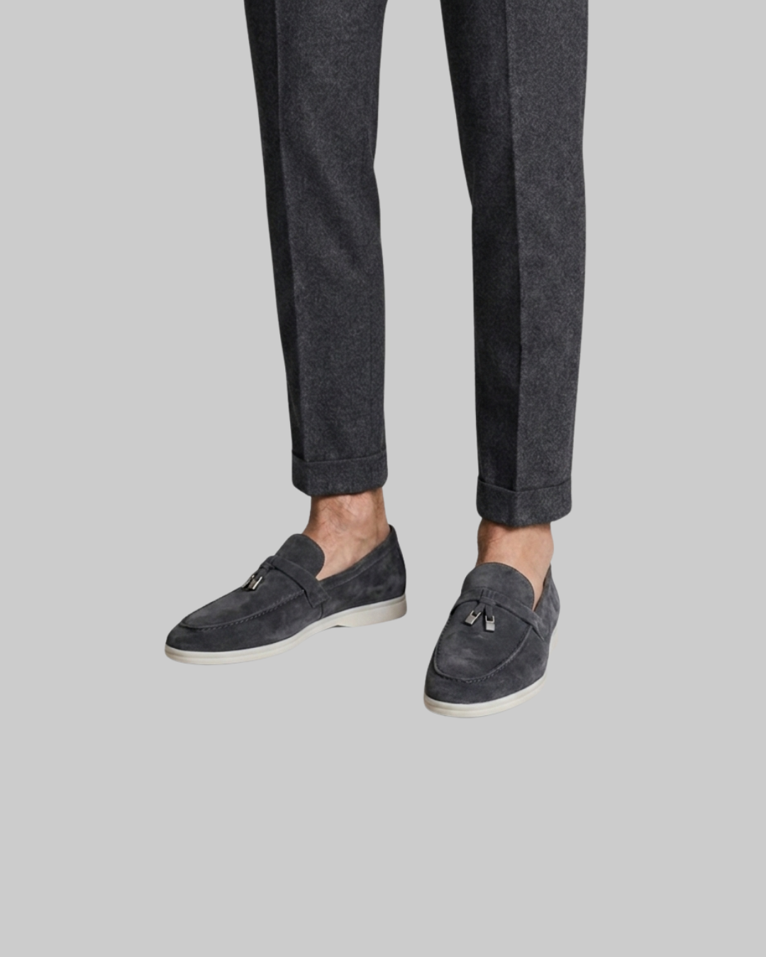 A person wearing dark charcoal gray trousers and matching dark gray suede tassel loafers with white soles, shown from the calves down against a solid light gray background.