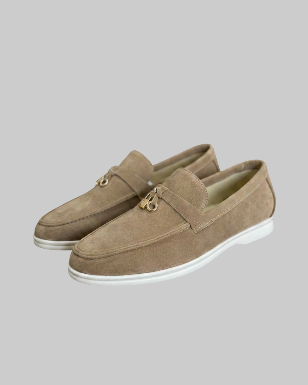 A pair of men's suede loafers in a light brown/tan color, featuring a small gold-tone metal ring detail on the vamp and a white sole, set against a solid gray background.