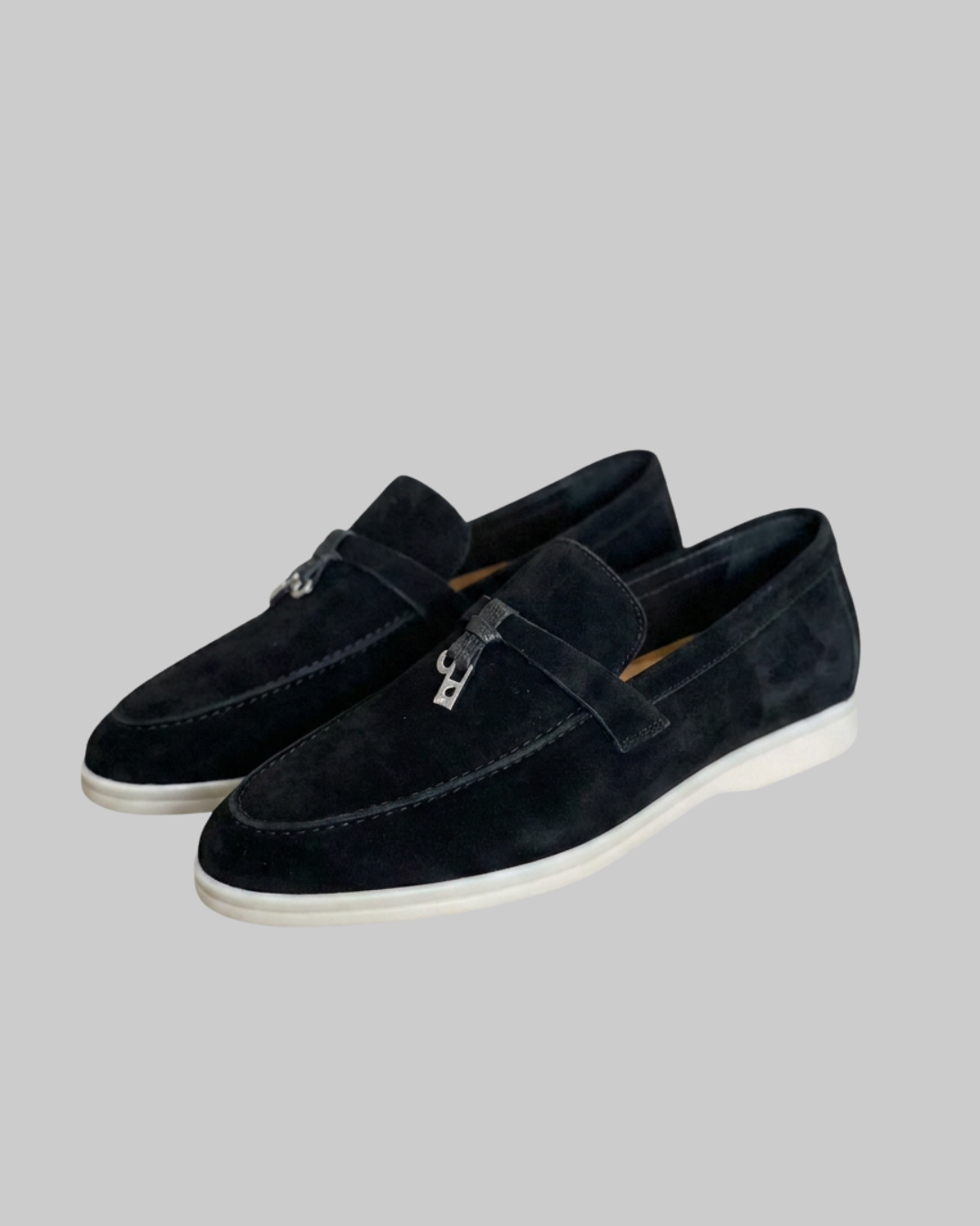 A pair of men's black suede loafers with silver-tone metal hardware and white soles, displayed on a solid gray background.