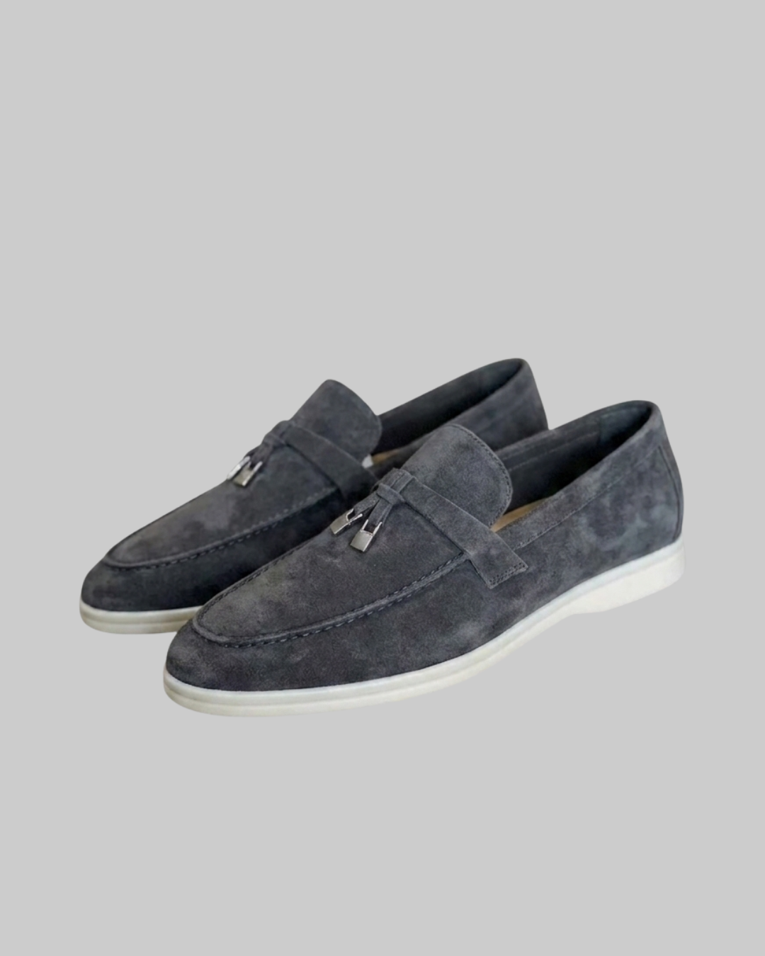 A pair of men's gray suede loafers with silver-tone metal tassel details and white soles, shown against a solid gray background.