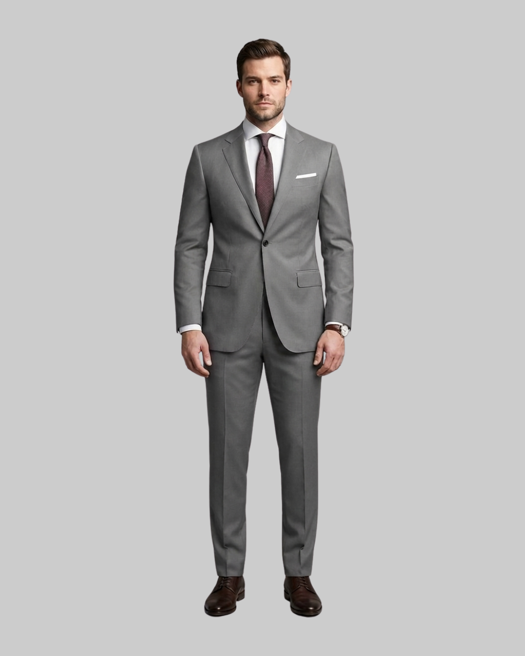 A man modeling a classic gray two-piece suit with a white dress shirt, a dark burgundy tie, and brown dress shoes, set against a solid gray studio background.