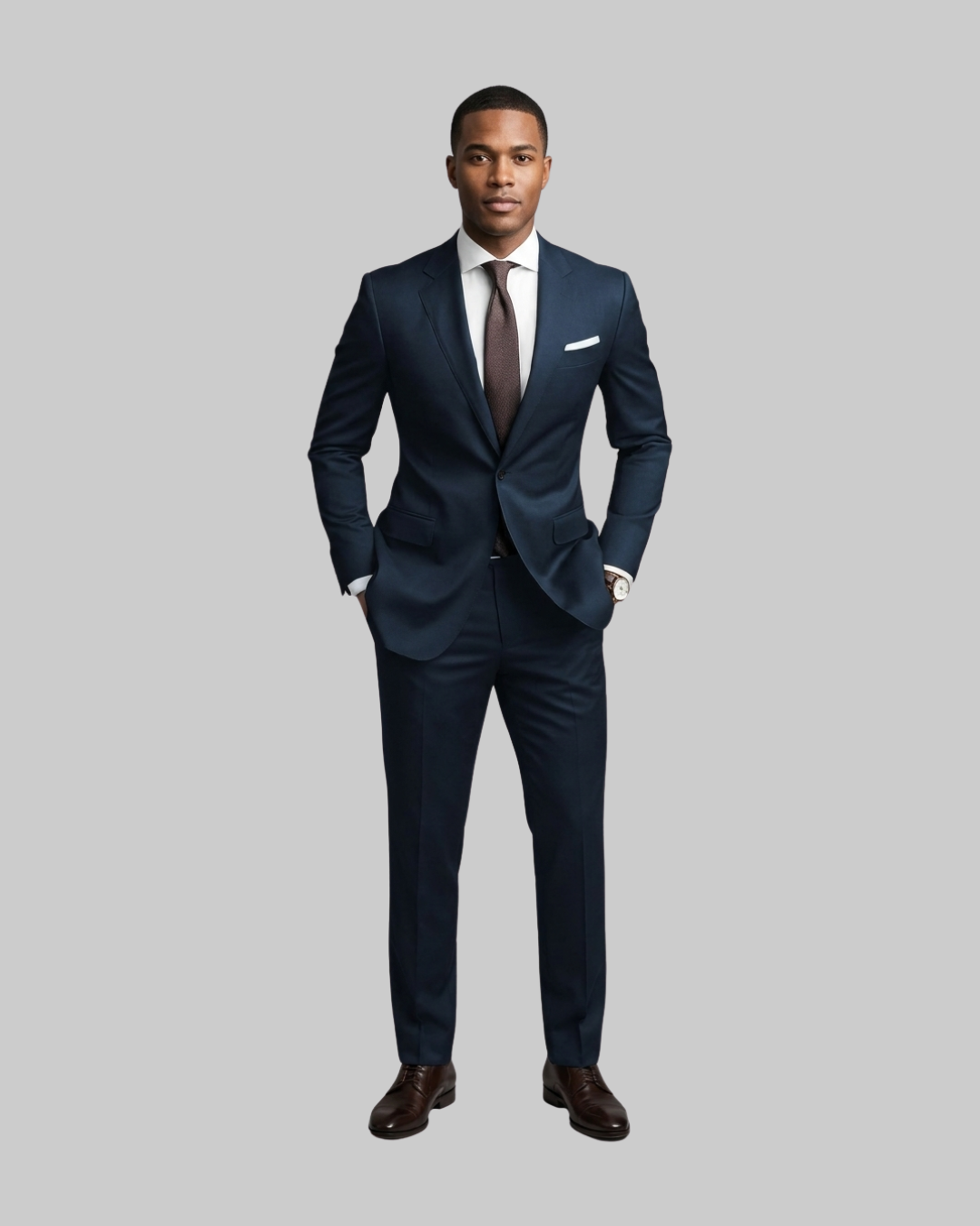 A man modeling a navy blue two-piece suit with a white dress shirt, a dark brown tie, a white pocket square, and dark brown dress shoes, set against a solid gray background.