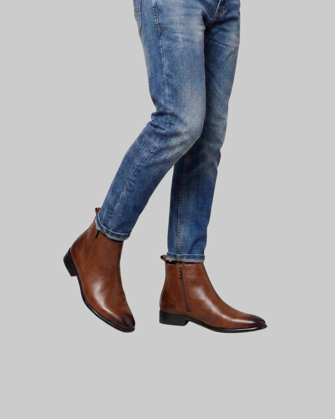 A person wearing blue jeans and brown leather ankle boots with side zippers, shown from the knees down against a plain light gray background.