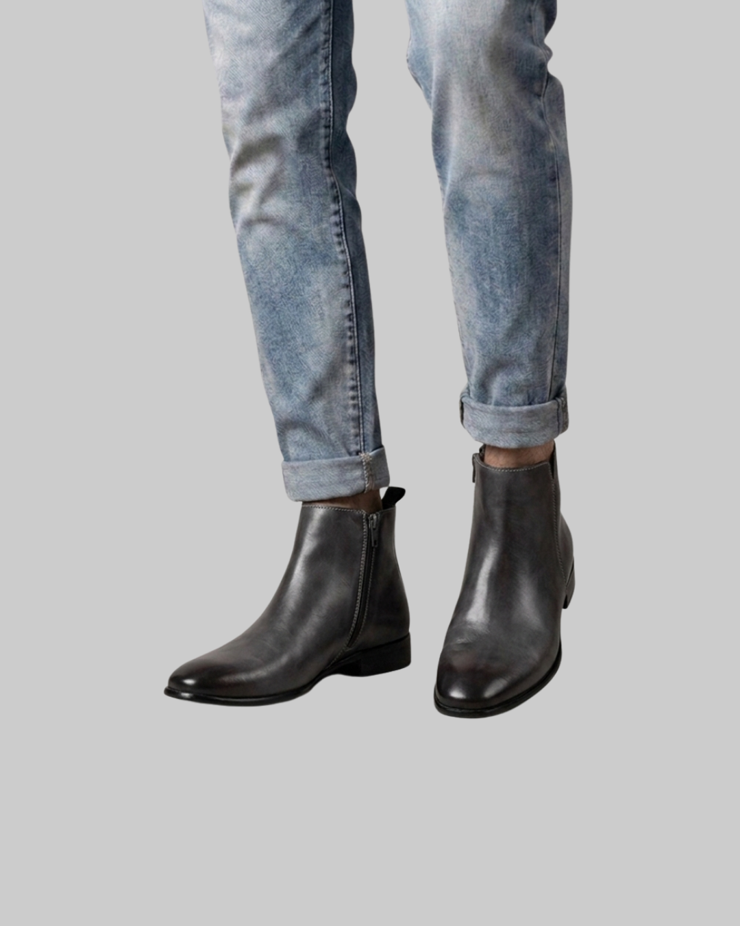 A person wearing black leather ankle boots with light-wash blue jeans, shown from the calves down against a solid gray background.