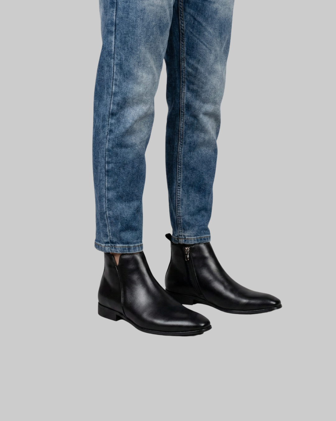 Black leather ankle boots with side zippers, shown on a model wearing blue jeans against a solid gray background.