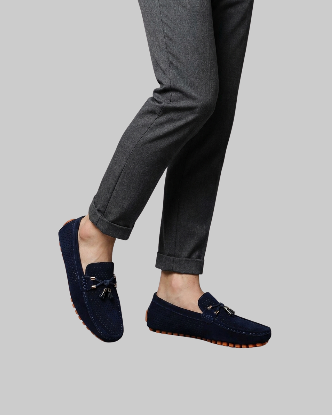 A person wearing dark gray cuffed trousers and navy blue suede loafers with gold-tone tassel details, shown against a solid light gray background.