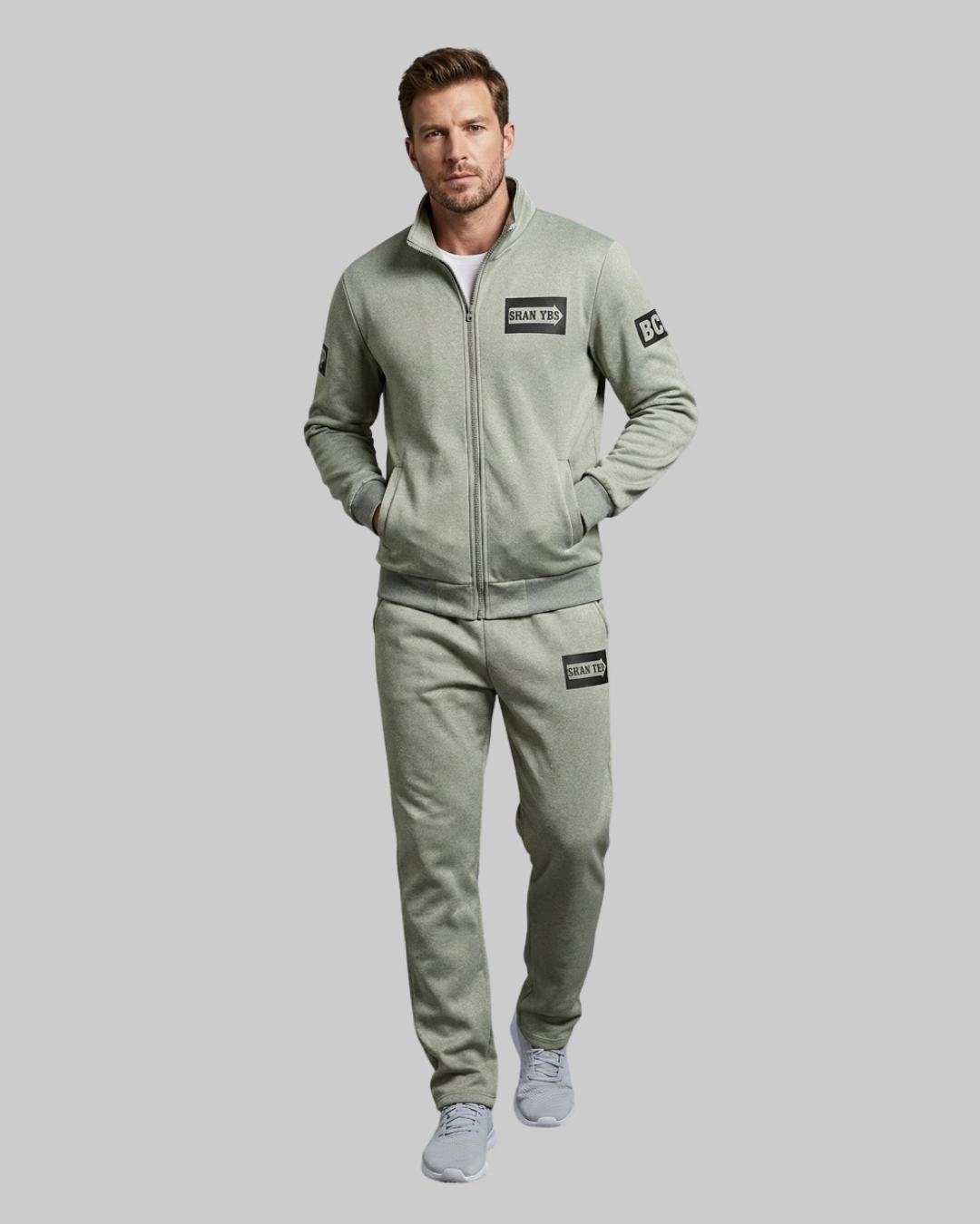 A man modeling a matching light sage green zip-up sweatshirt and jogger pants set with black brand patches, photographed against a solid gray studio background.