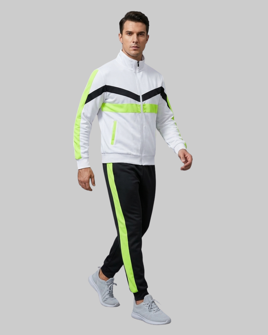 A man modeling a white athletic zip-up jacket with black and neon green stripe details, paired with black track pants featuring a neon green side stripe and light gray sneakers, set against a solid gray background.