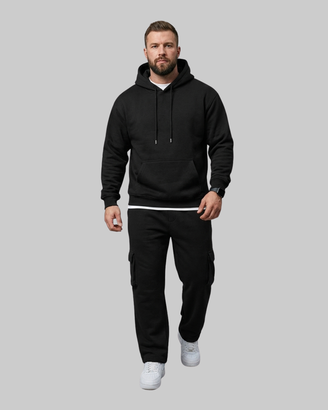A man wearing a black pullover hoodie with black drawstrings, a black sweatshirt, and black cargo pants with white sneakers, standing against a solid gray background.