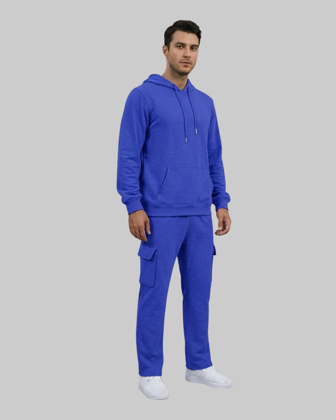 A man modeling a matching royal blue sweatshirt and cargo pants set, paired with white sneakers, against a solid gray background.