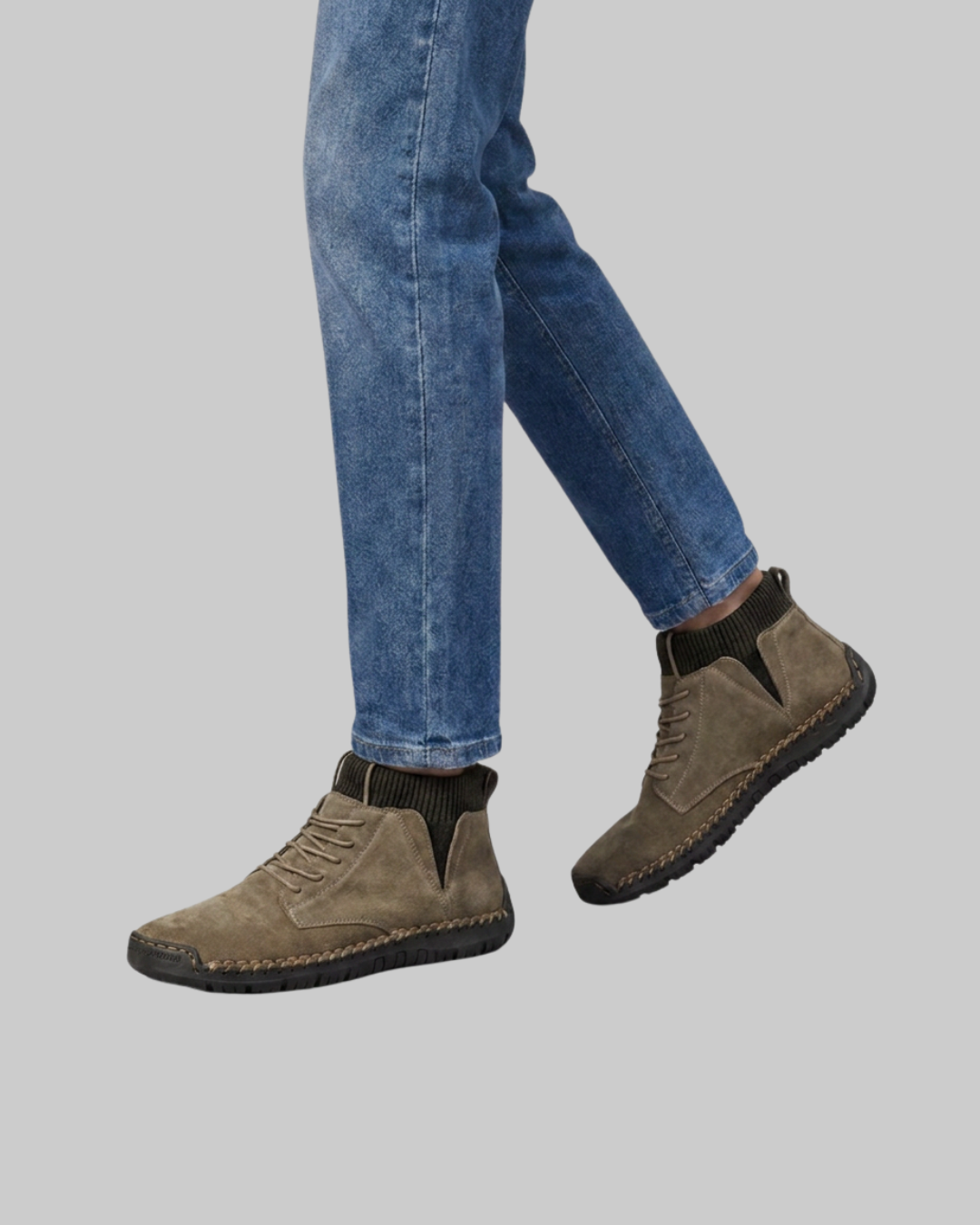A person wearing blue jeans and taupe-brown suede ankle boots with dark elastic side panels, shown against a solid gray background.