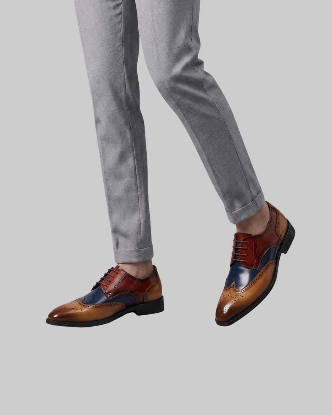 A person wearing gray trousers and brown and navy color-blocked wingtip oxford shoes, shown from the waist down against a solid gray background.