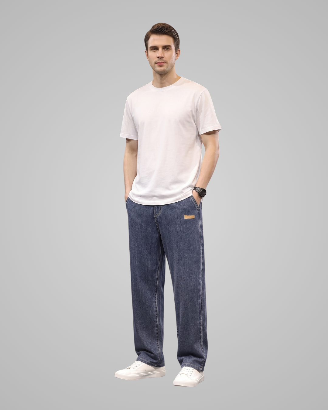 A man modeling a white crew-neck t-shirt and dark blue relaxed-fit jeans with white sneakers, standing against a solid gray background.